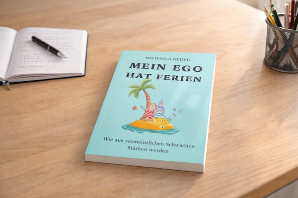 Buch ‚Mein Ego hat Ferien‘ auf einem Schreibtisch neben einem geöffneten Notizbuch und Stiften.