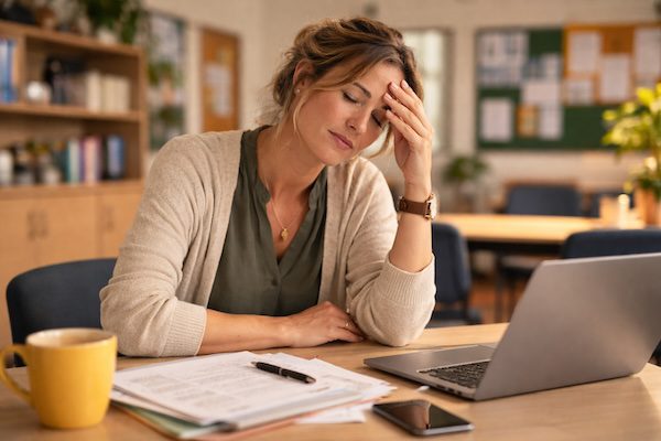 Person sitzt am Schreibtisch, wirkt erschöpft und hält die Hand an die Stirn, umgeben von Unterlagen und Laptop.